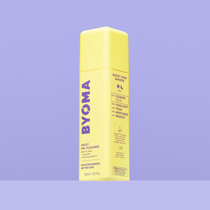 Une bouteille jaune de Byoma Milky Oil Cleanser, vue inclinée montrant les étapes de routine « Boost Your Barrier » sur le panneau latéral, sur fond violet.