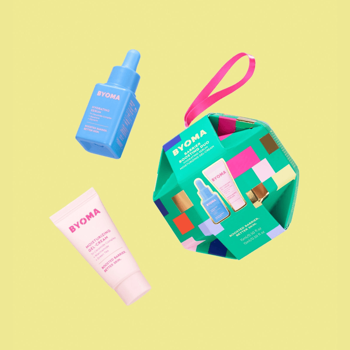 Un coffret cadeau vert multicolore en forme d'ornement intitulé « BYOMA Barrier Boosting Duo », comprenant un tube rose de gel-crème hydratant et une bouteille bleue de sérum hydratant. Les éléments sont affichés flottants sur un fond jaune.